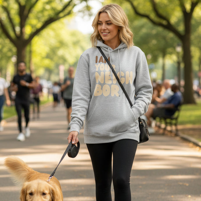 Woman walking through a city park with her dog wearing the APWT Gift Shop's I Am Your Neighbor Premium Unisex Hoodie from the Day in the Life Collection and the Premium Basics Collection in Grey