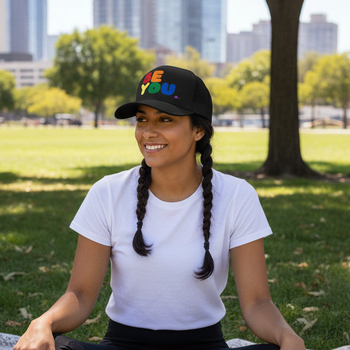 Woman sitting in the park in the grass wearing the APWT Gift Shop's Rainbow Be You Trucker Hat from the Pride in the APWT Gift Shop Collection and the Be You Collection in Black