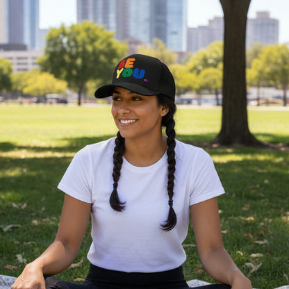 Woman sitting in the park in the grass wearing the APWT Gift Shop's Rainbow Be You Trucker Hat from the Pride in the APWT Gift Shop Collection and the Be You Collection in Black