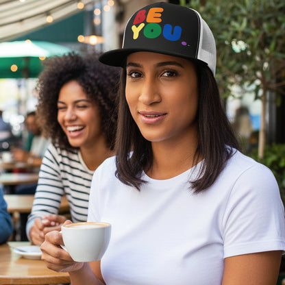 Woman sitting at an outdoor cafe wearing the APWT Gift Shop's Rainbow Be You Trucker Hat from the Pride in the APWT Gift Shop Collection and the Be You Collection in black and white