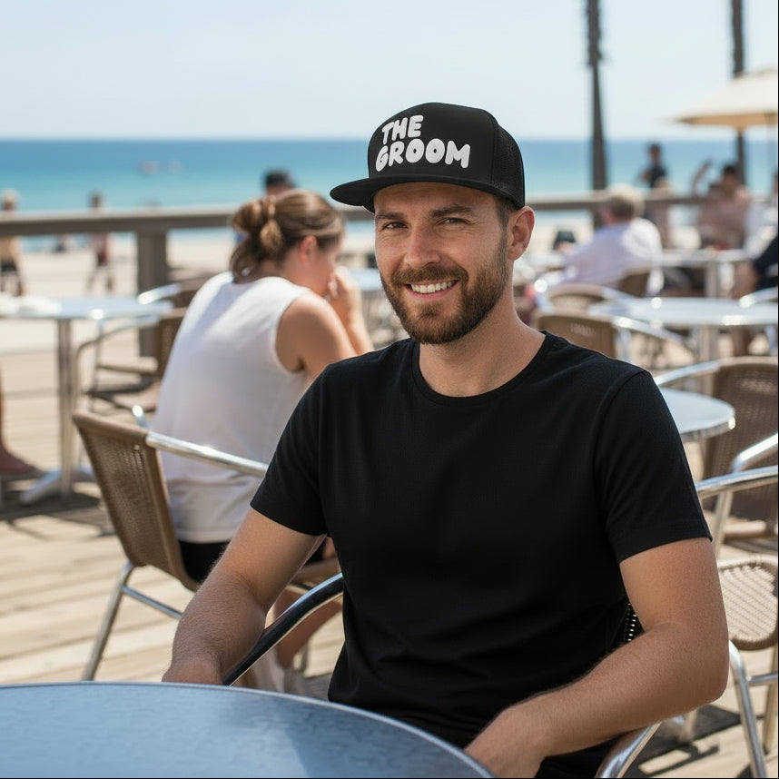 Man at an outdoor cafe wearing the  APWT Gift's Shop Groom Hat featuring a charcoal grey hat that has 'The Groom' featured in white letters