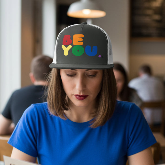 Woman sitting in a cafe wearing the APWT Gift Shop's Rainbow Be You Trucker Hat from the Pride in the APWT Gift Shop Collection and the Be You Collection in charcoal grey and white