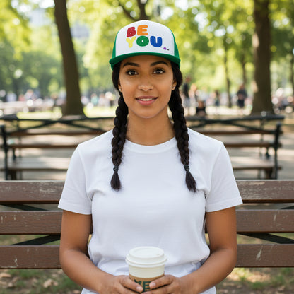 Woman sitting on a bench in the park wearing the APWT Gift Shop's Rainbow Be You Trucker Hat from the Pride in the APWT Gift Shop Collection and the Be You Collection in green and white