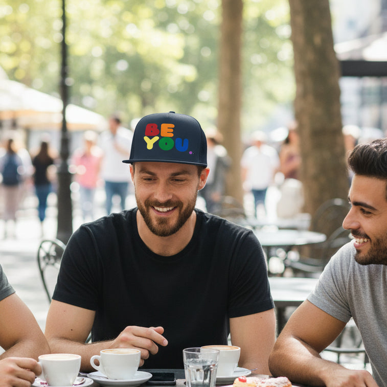 Man sitting at an outdoor cafe wearing the APWT Gift Shop's Rainbow Be You Trucker Hat from the Pride in the APWT Gift Shop Collection and the Be You Collection in Navy Blue