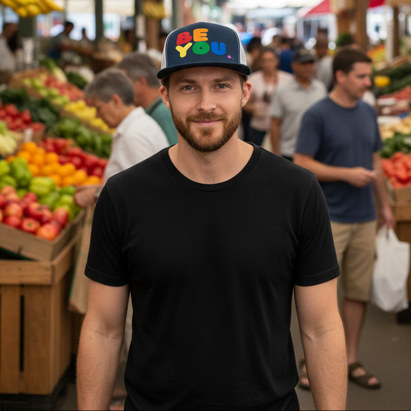 Man standing in a farmer's market wearing the APWT Gift Shop's Rainbow Be You Trucker Hat from the Pride in the APWT Gift Shop Collection and the Be You Collection in Navy blue and white
