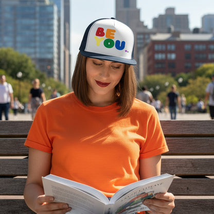 Woman sitting sitting on a park bench reading a book wearing the APWT Gift Shop's Rainbow Be You Trucker Hat from the Pride in the APWT Gift Shop Collection and the Be You Collection in navy blue and white