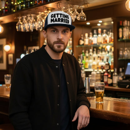 Man standing in a bar wearing the APWT Gift Shop Getting Married Trucker Hat in Navy Blue and White