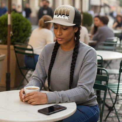 Woman sitting at a cafe with a cup of coffee wearing the APWT Gift Shop's Shades in Black Trucker Hat in Silver and Black