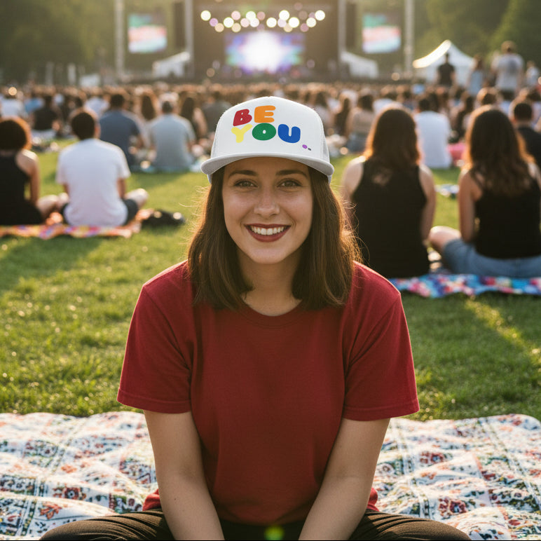 Woman sitting at an outdoor concert wearing the APWT Gift Shop's Rainbow Be You Trucker Hat from the Pride in the APWT Gift Shop Collection and the Be You Collection in white