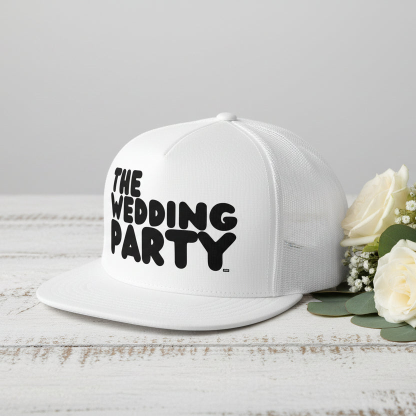 The APWT Gift Shop's The Wedding Party Trucker Hat from the APWT Wedding Shop in White sitting on a  white wooden table next to flowers