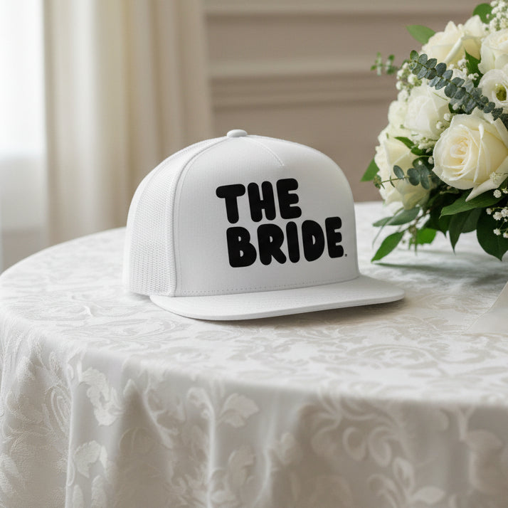 The Bride Trucker Hat from the APWT Gift Shop in white sitting on a table
