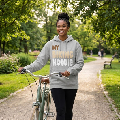 Woman walking her bike through a park wearing the APWT Gift Shop's My Biking Hoodie Premium Unisex Hoodie from the Day in the Life Collection and the Premium Basics Collection in grey
