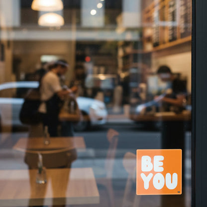 The APWT Gift Shop's Be You in Orange Vinyl Sticker stuck to a the outside of a glass window of a cafe