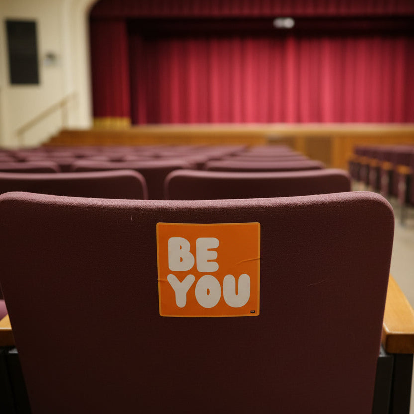 The APWT Gift Shop's Be You in Orange Vinyl Sticker stuck to a the back of a chair in an auditorium