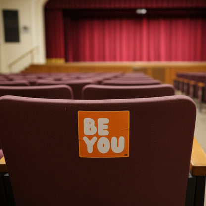 The APWT Gift Shop's Be You in Orange Vinyl Sticker stuck to a the back of a chair in an auditorium
