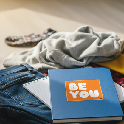 The APWT Gift Shop's Be You in Orange Vinyl Sticker stuck to the front of a book laying in a pile of clothes