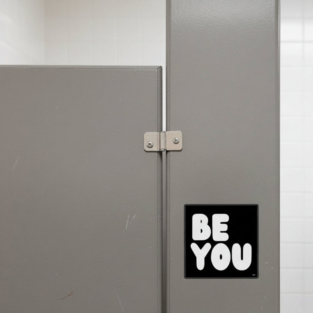 The APWT Gift Shop's Be You in Black and White Vinyl Sticker from the Be You Collection stuck outside of a stall in a public bathroom