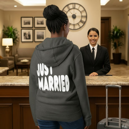 Woman standing at a hotel desk wearing the APWT Gift Shop's Just Married Premium Unisex Zip-Up Hoodie in Charcoal Grey from the APWT Wedding Shop