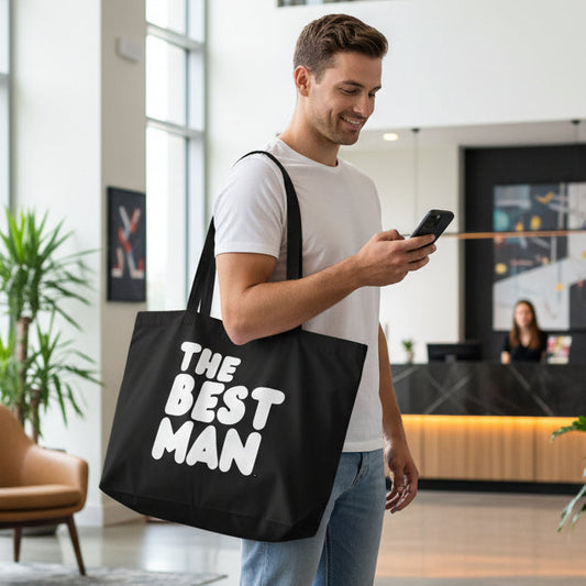 Man standing in a hotel lobby carrying the APWT Gift Shop's the Best Man Jumbo Black Tote Bag from the APWT Wedding Shop