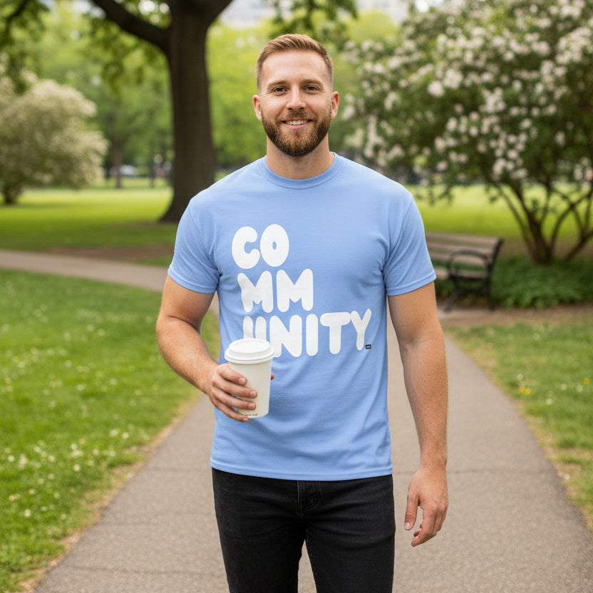 Man standing in the park wearing the APWT Gift Shop's A Community in Sky Blue Unisex T-Shirt from the Community Collection