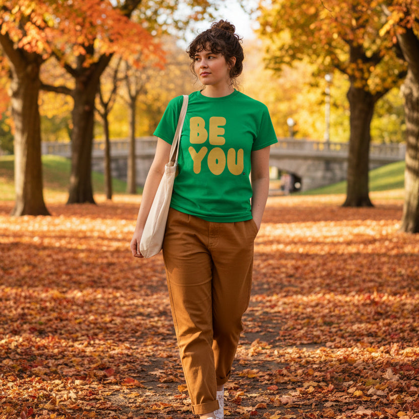 Woman walking through the park on an autumn day wearing the APWT Gift Shop's Be You in Green and Yellow Unisex T-Shirt from the Be You Collection