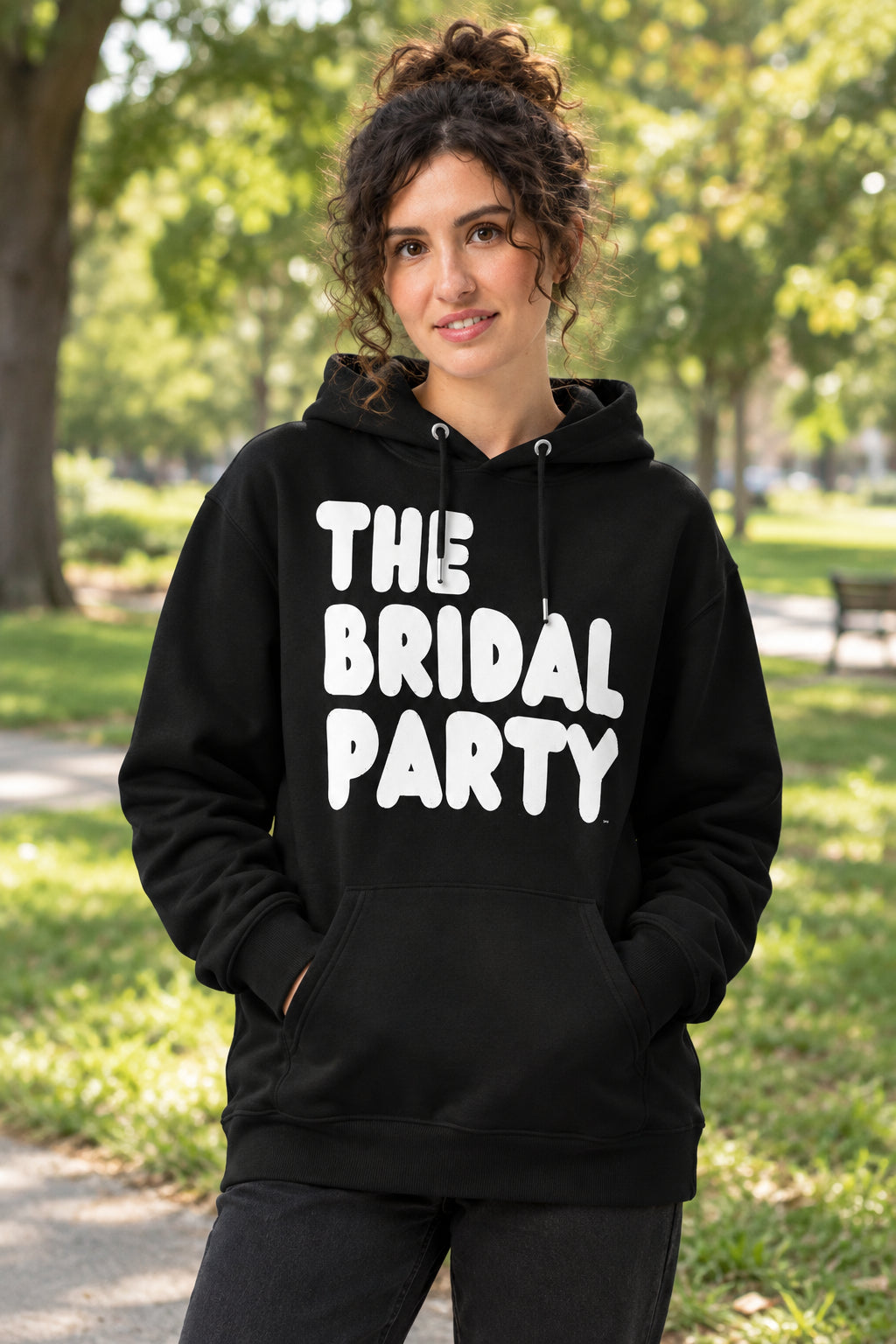 Woman standing in a park wearing the APWT Gift Shop's The Bridal Party Premium Unisex Hoodie from the APWT Wedding Shop in Black