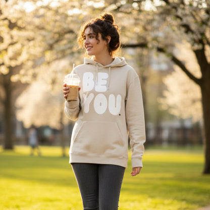 Woman walking through a city park wearing the APWT Gift Shop's Be You in Beige and White Premium Unisex Hoodie from the Be You Collection and the Premium Basics Collection
