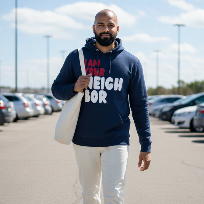 Man walking through a parking lot wearing the APWT Gift Shop's I Am Your Neighbor in Turmoil Premium Unisex Hoodie from the Day in the Life Collection and the Premium Basics Collection