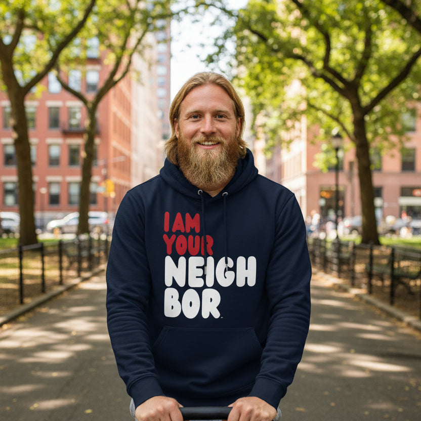 Man pushing a baby stroller through a city park wearing the APWT Gift Shop's I Am Your Neighbor in Turmoil Premium Unisex Hoodie from the Day in the Life Collection and the Premium Basics Collection