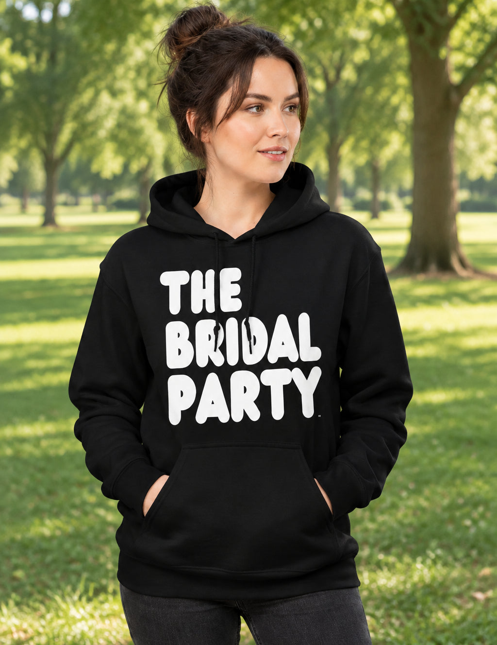Woman standing in a park wearing the APWT Gift Shop's The Bridal Party Unisex Hoodie from the APWT Wedding Shop in Black