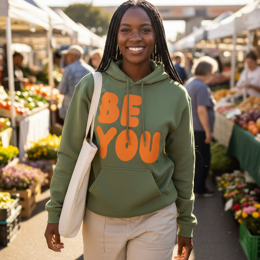 Woman walking through a farmer's market wearing the APWT Gift Shop's Be You in Olive Green and Orange Unisex Hoodie from the Be You Collection