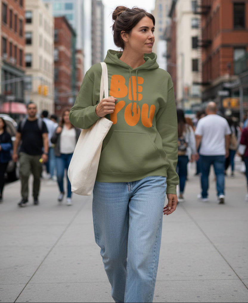 Woman walking on a city sidewalk wearing the APWT Gift Shop's Be You in Olive Green and Orange Unisex Hoodie from the Be You Collection