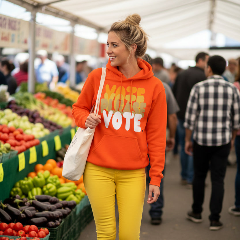 Woman walking through a farmer's market wearing the APWT Gift Shop's Please Vote in Safety Cone Orange Unisex Hoodie from the APWT Vote Shop