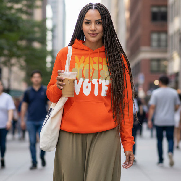 Woman walking down a city street wearing the APWT Gift Shop's Please Vote in Safety Cone Orange Unisex Hoodie from the APWT Vote Shop
