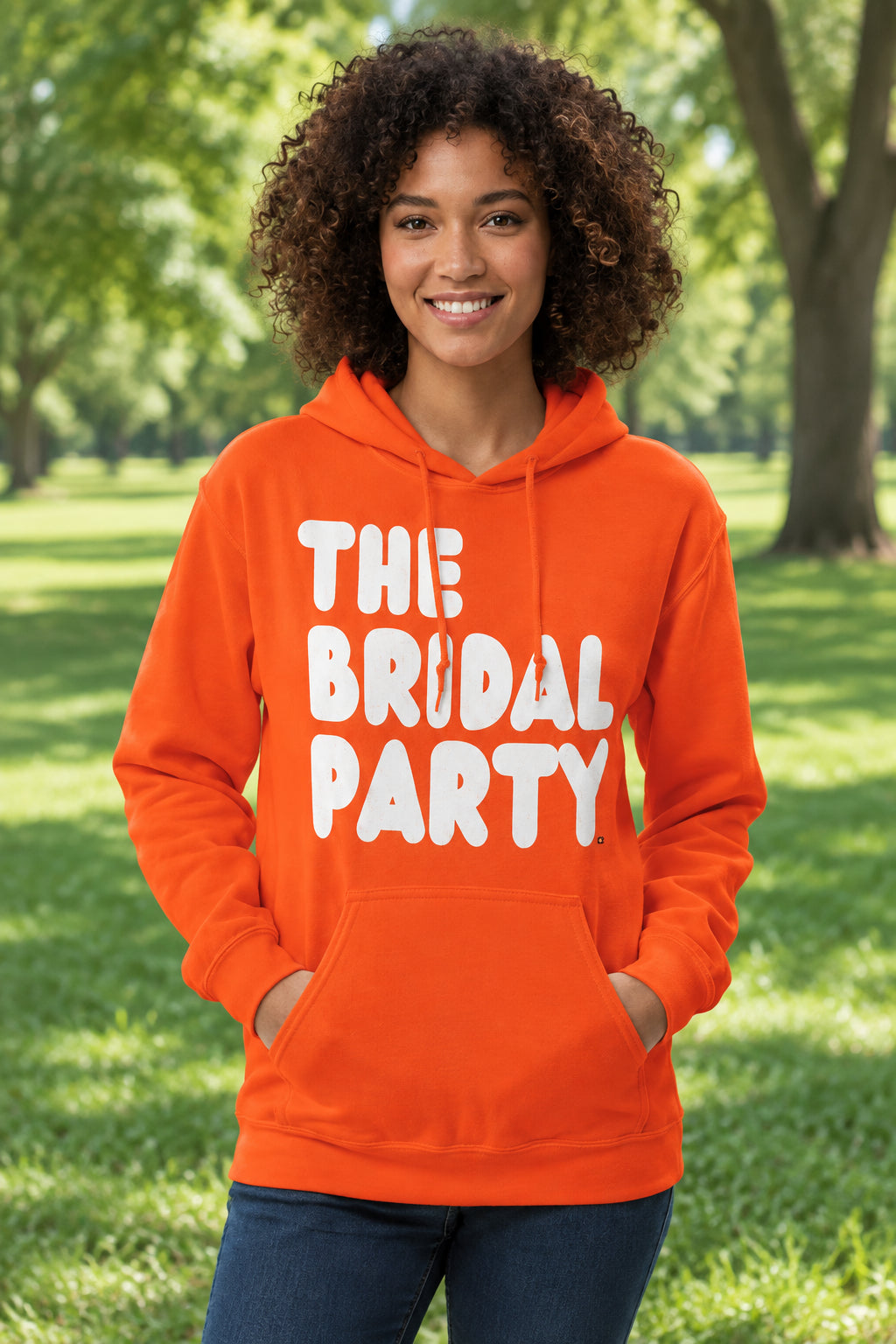 Woman standing in a park wearing the APWT Gift Shop's The Bridal Party Unisex Hoodie from the APWT Wedding Shop in Orange