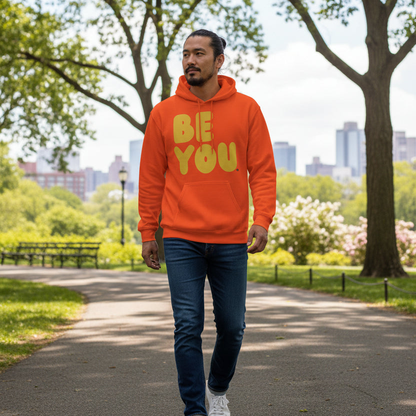 Man walking through a city park wearing the APWT Gift Shop's Be You in Orange and Yellow Unisex Hoodie from the Be You Collection