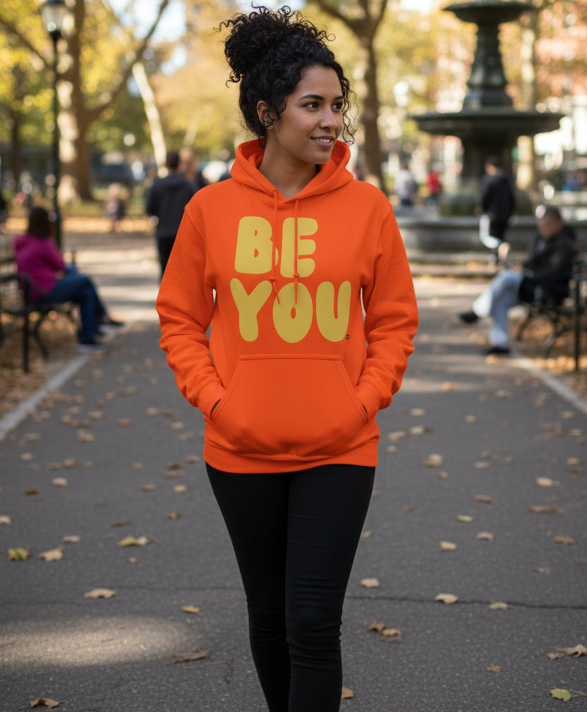 Woman walking through a city park wearing the APWT Gift Shop's Be You in Orange and Yellow Unisex Hoodie from the Be You Collection