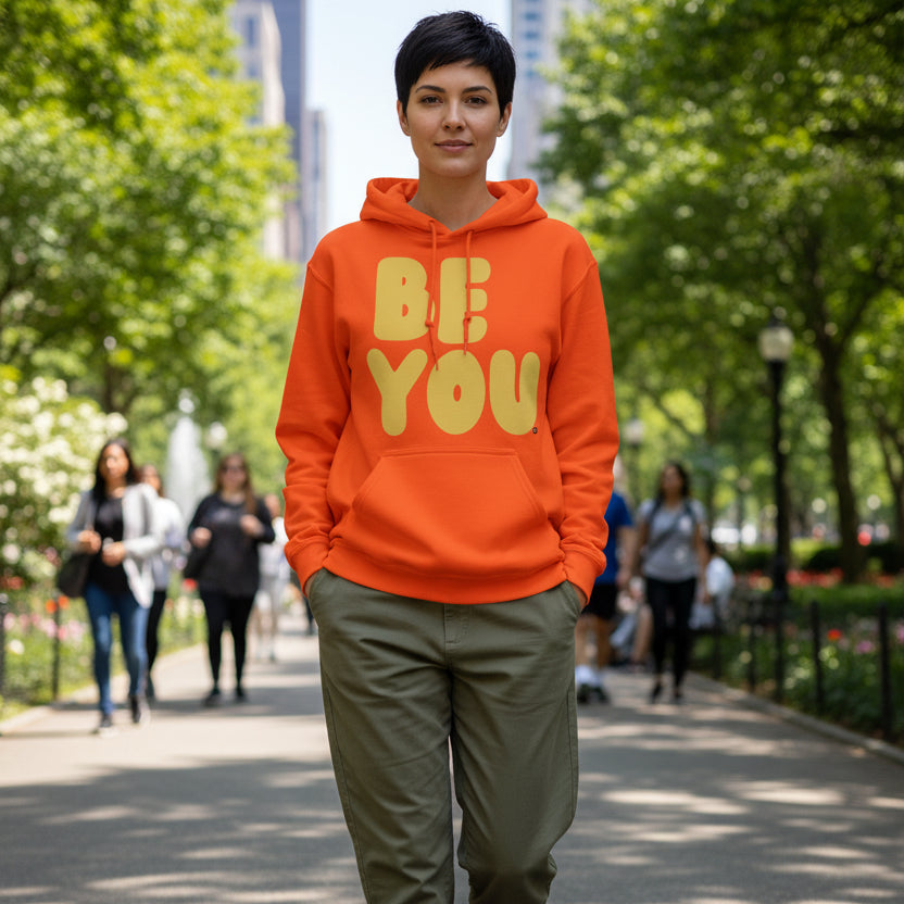 Woman walking through a city park wearing the APWT Gift Shop's Be You in Orange and Yellow Unisex Hoodie from the Be You Collection