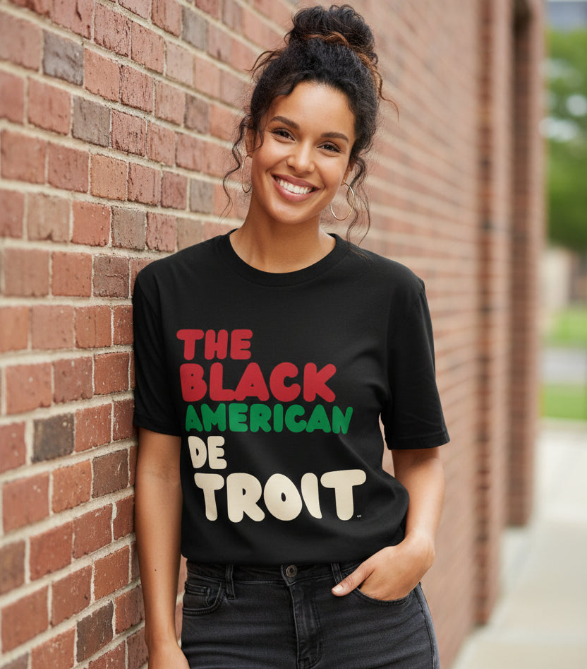Woman leaning against a brick wall wearing the APWT Gift Shop's the Black American Detroit Premium Unisex T-shirt from the Black Collection