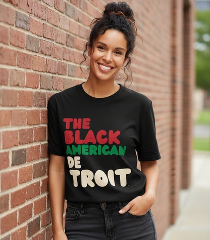 Woman leaning against a brick wall wearing the APWT Gift Shop's the Black American Detroit Premium Unisex T-shirt from the Black Collection