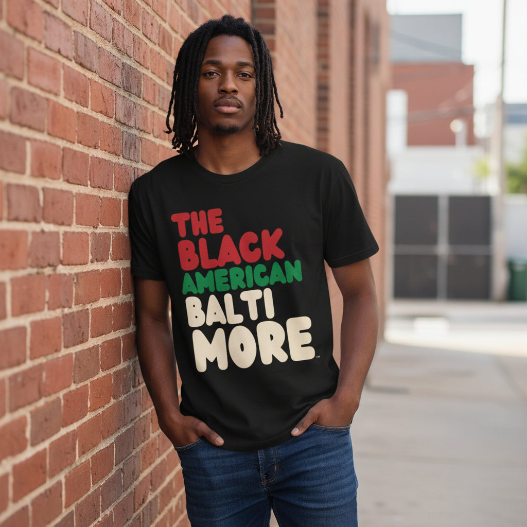 Man leaning against a brick wall wearing the APWT Gift Shop's the Black American Baltimore Premium Unisex T-Shirt from the Black Collection