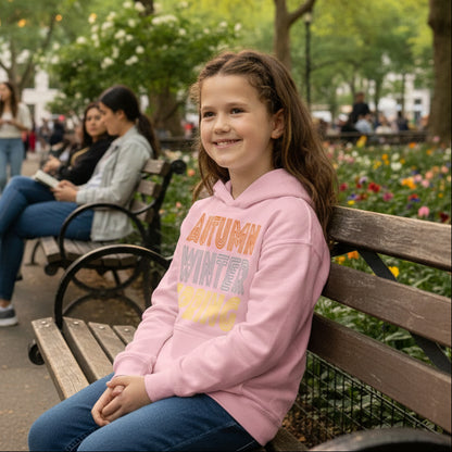 Girl sitting in a city park wearing The APWT Gift Shop's Fall into Autumn Kid's Unisex Hoodie in Pink from the APWT Kid's Shop